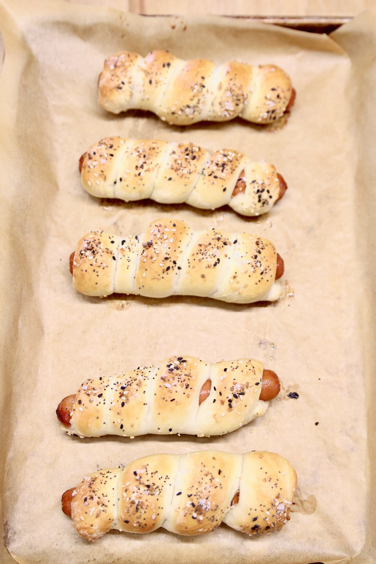5 baked pretzel dogs on a parchment lined baking sheet.