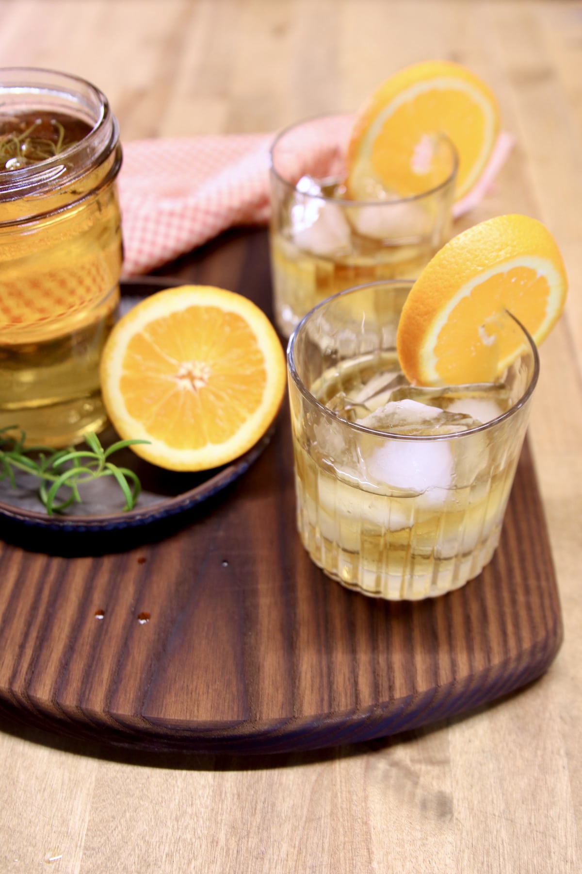 2 whiskey cocktails on a walnut board with half an orange, simple syrup.
