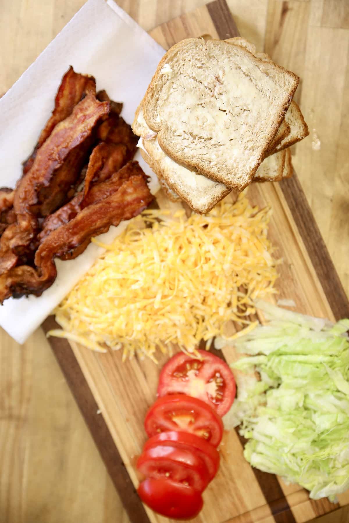 Cutting board with crisp bacon, buttered bread, shredded cheese. tomato, lettuce.