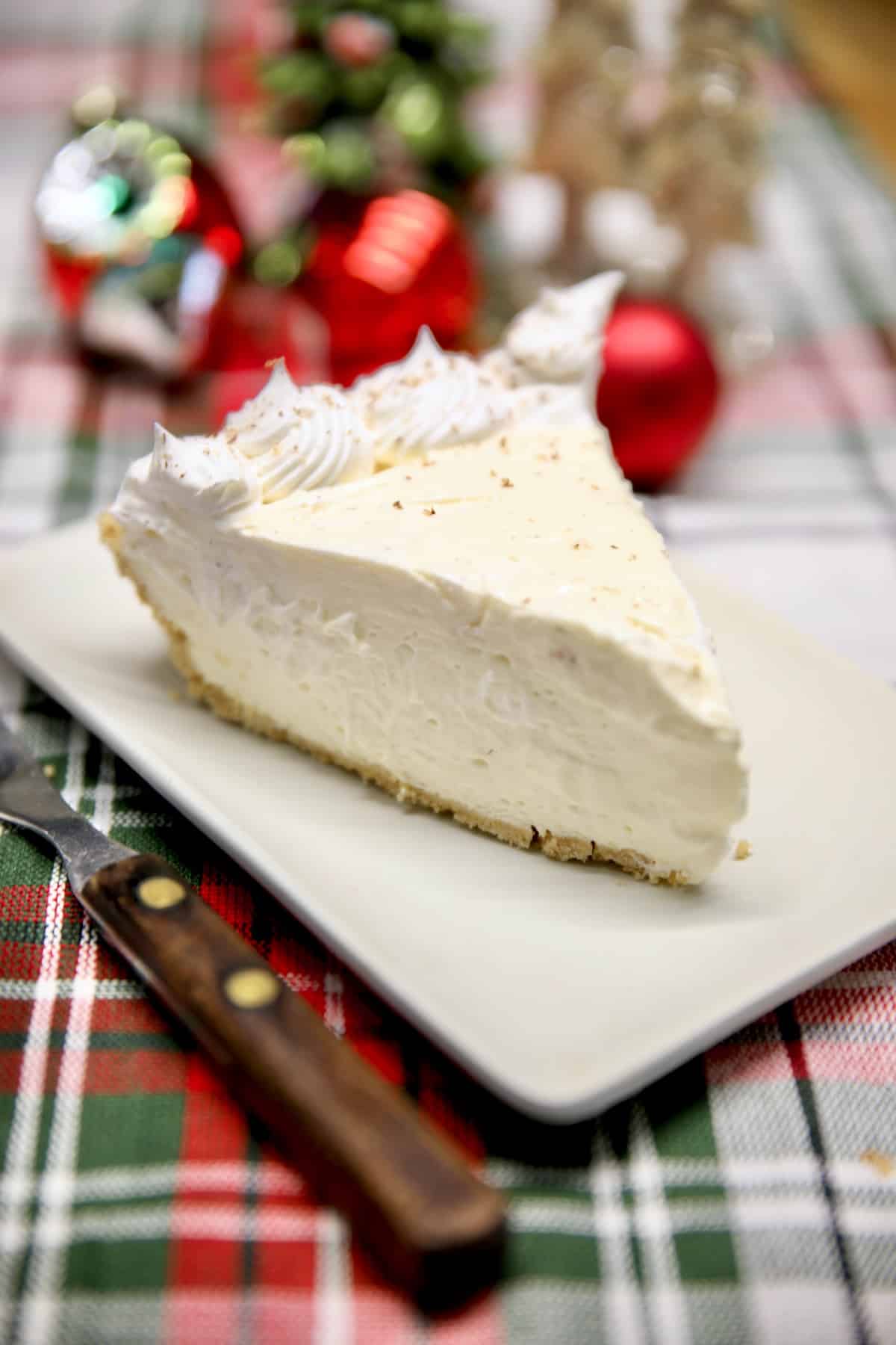 Slice of eggnog cheesecake on a plate, fork & Christmas decorations around the plate.