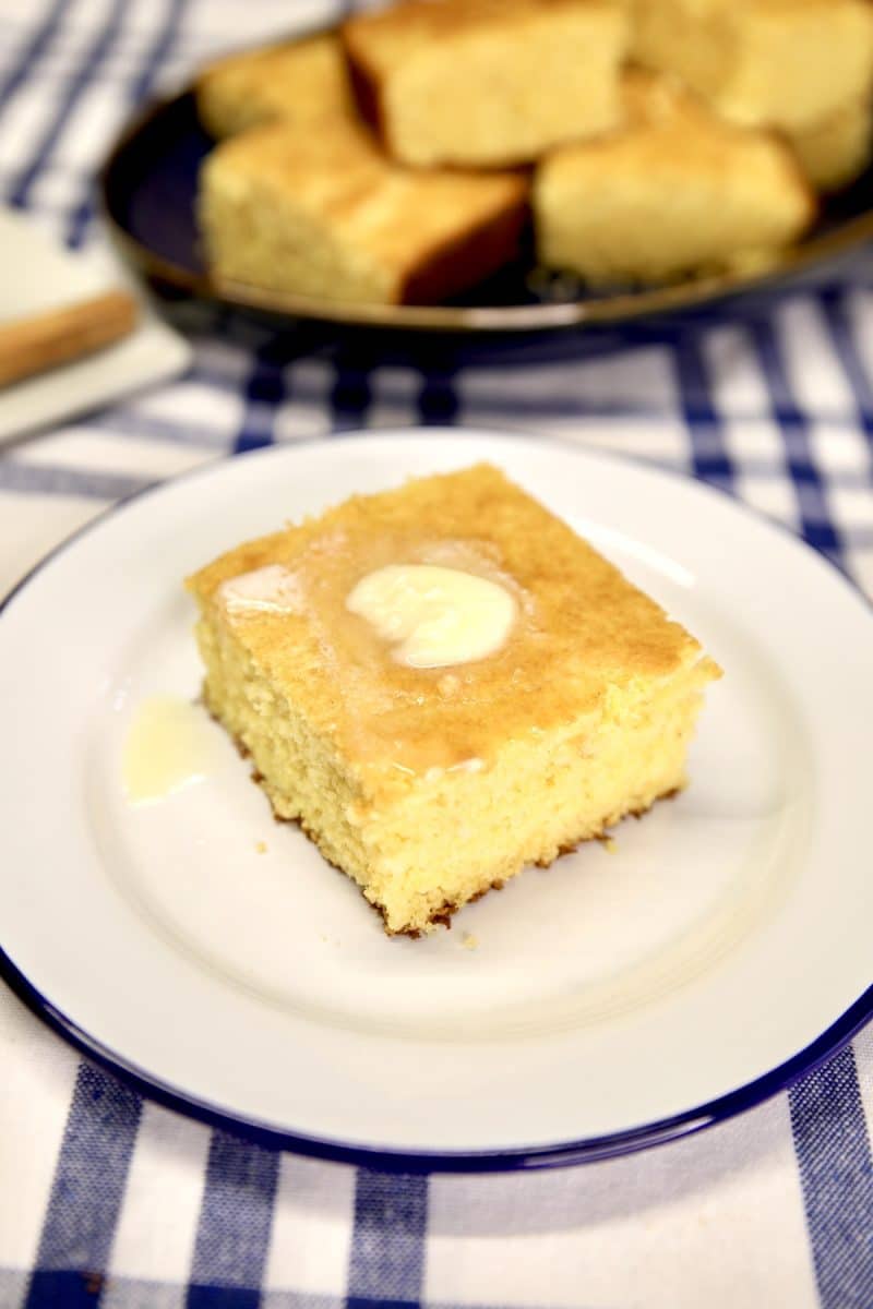 Cornbread slice on a plate with butter on top. Platter of cornbread in background.