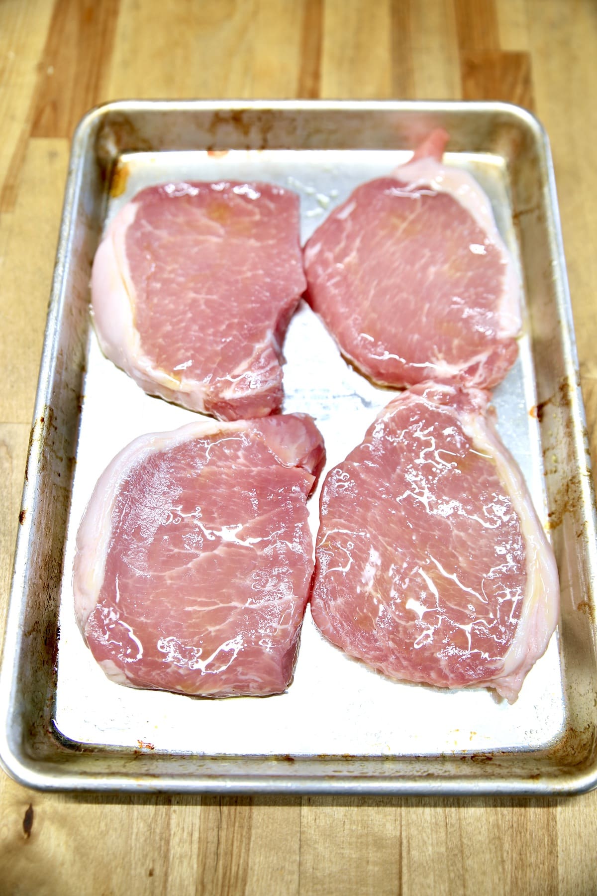 4 boneless raw pork chops coated with olive oil on a sheet pan.