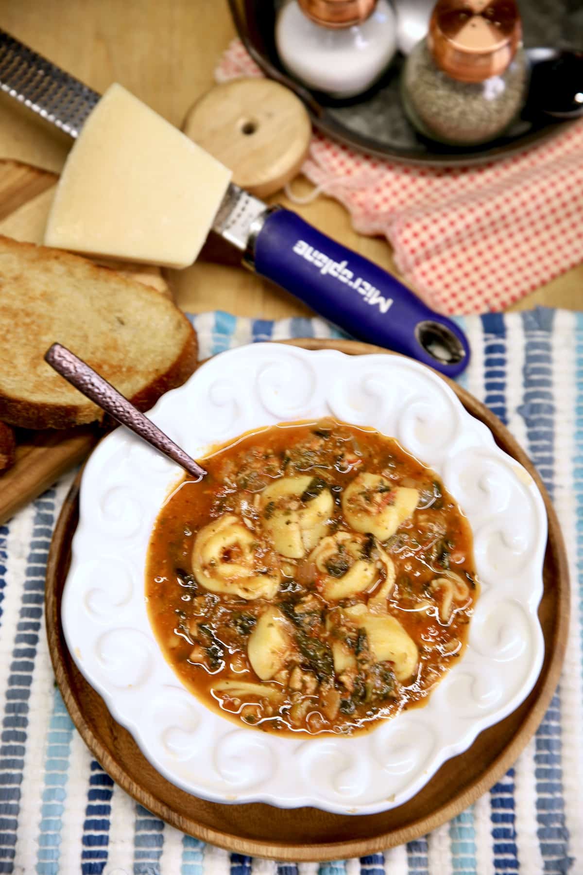 Bowl of tortellini soup, parmesan cheese with grater, bread. 