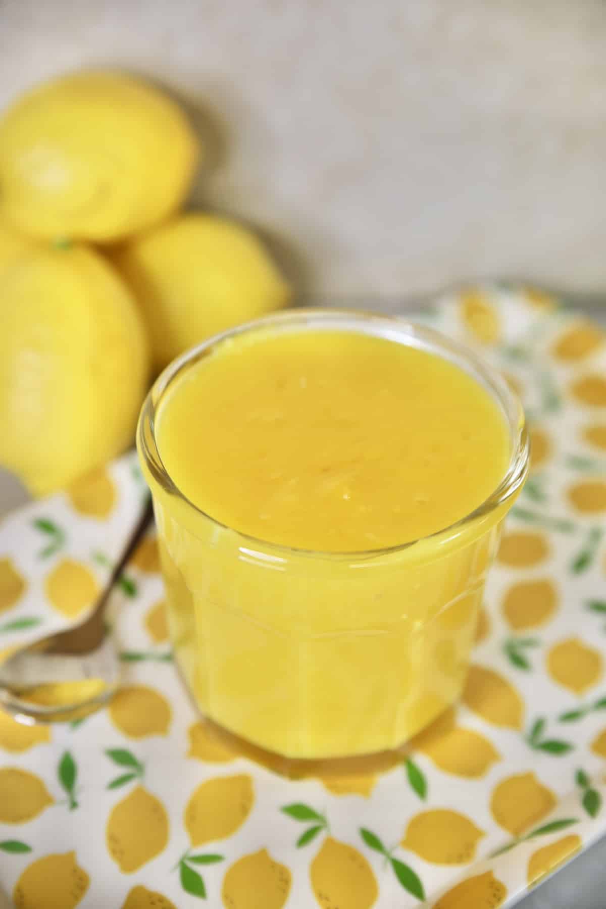 Jar of lemon curd on a lemon printed tray. Whole lemons in background. 