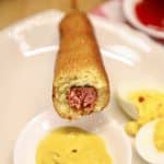 Smoked corn dog with bite out of it, dipping into mustard.