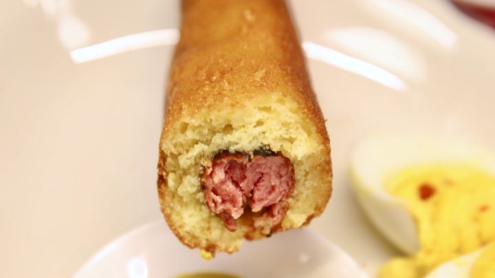 Smoked corn dog with bite out of it, dipping into mustard.