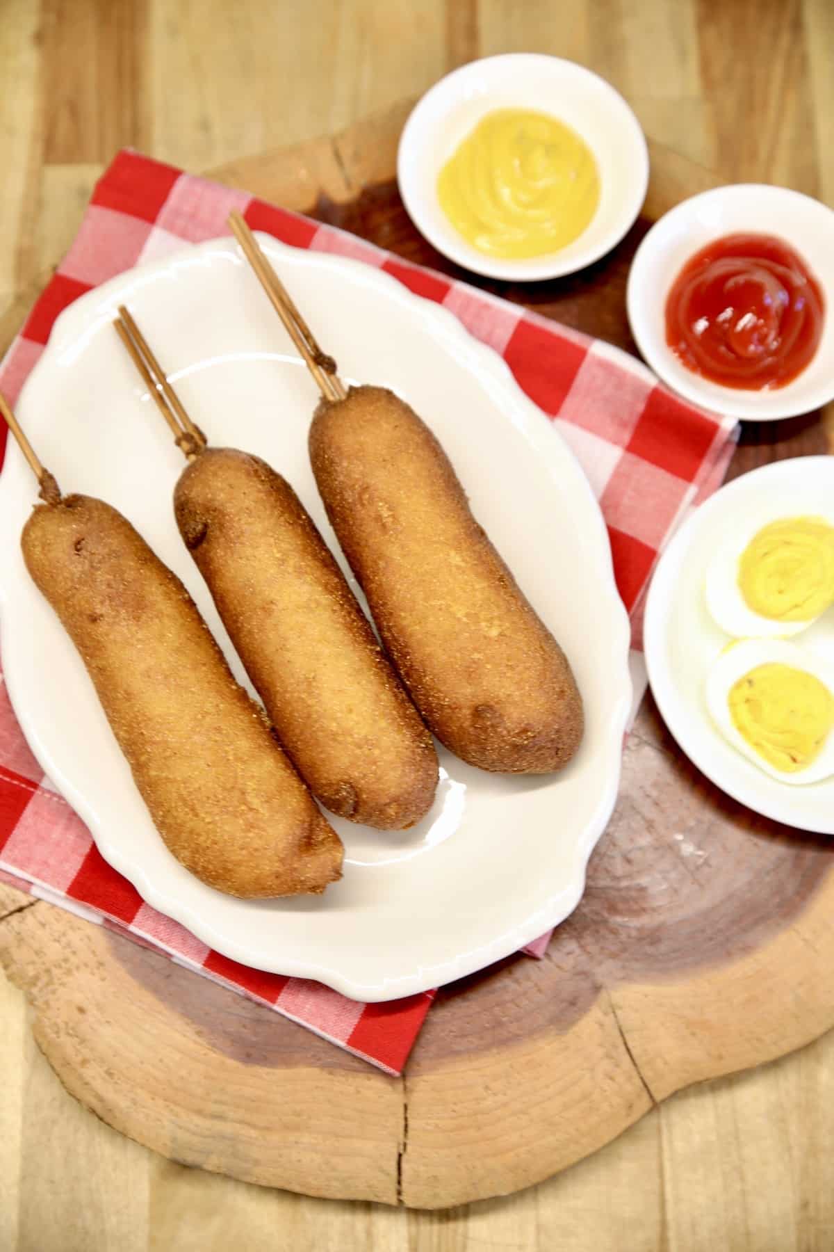 3 corn dogs on a platter, bowl of mustard, ketchup, deviled eggs.