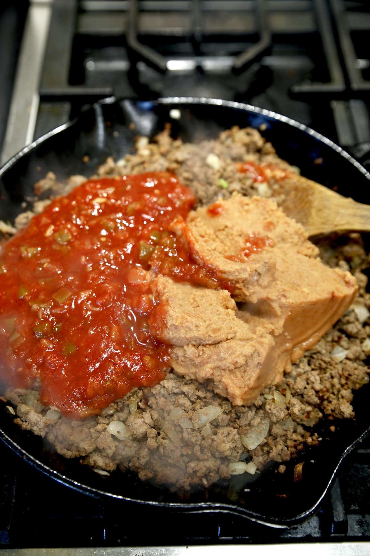 Adding picante sauce and refried beans to ground beef in a skillet.