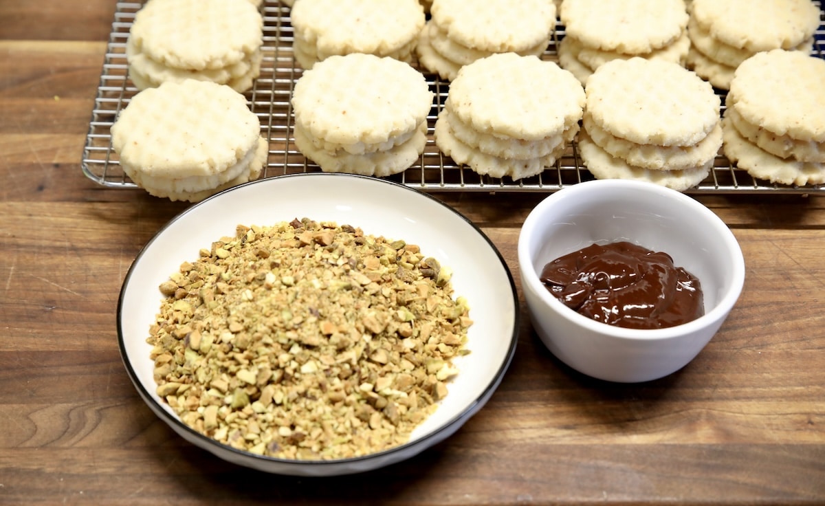 Cookies on a wire rack, chopped pistachios, melted chocolate in a bowl.