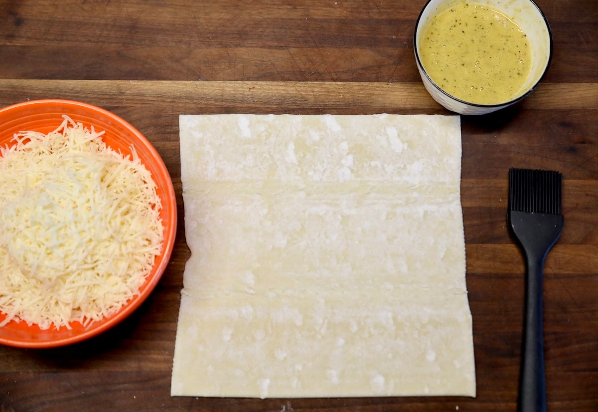 Puff pastry, shredded cheese and bowl of egg wash.