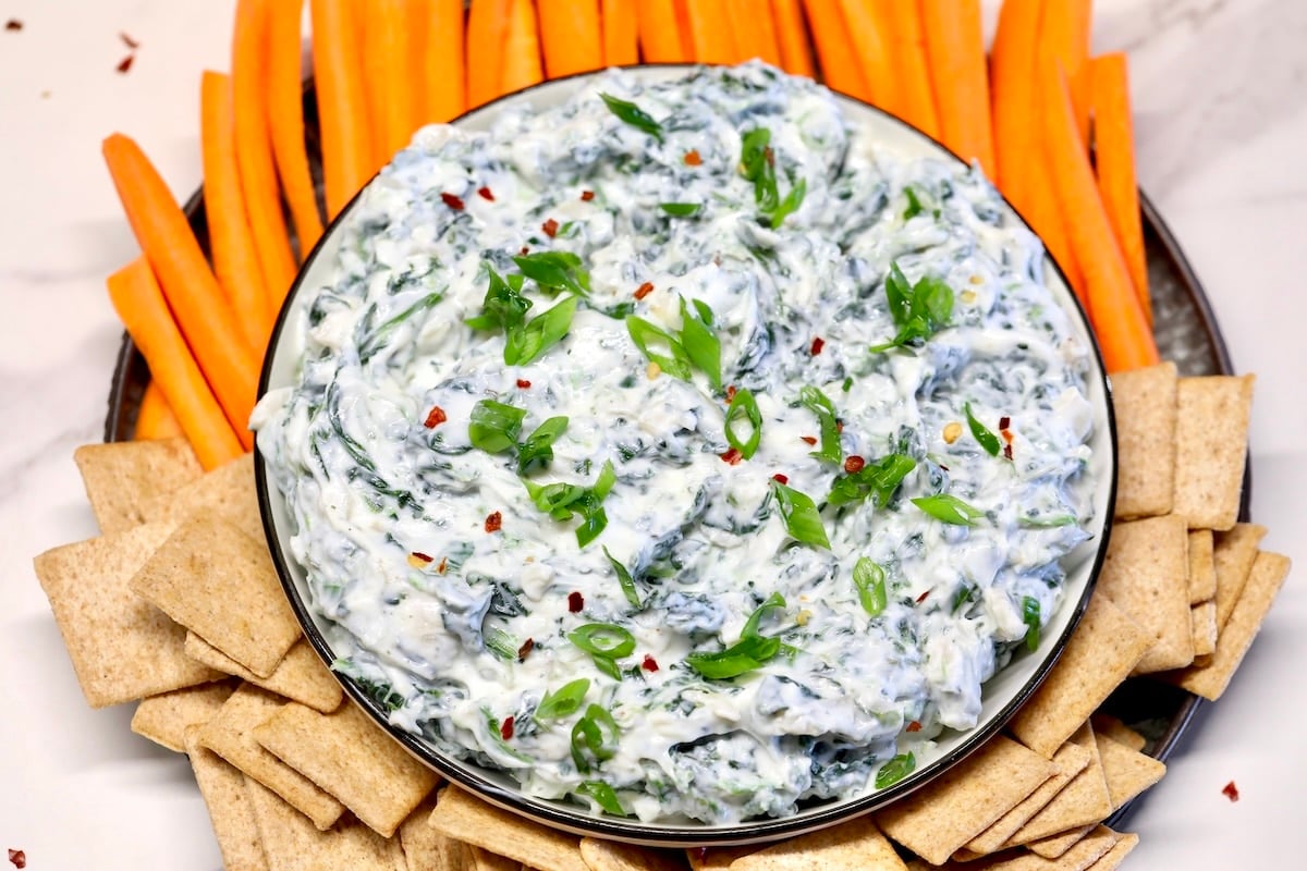 Bowl of spinach dip with carrot sticks and wheat crackers.