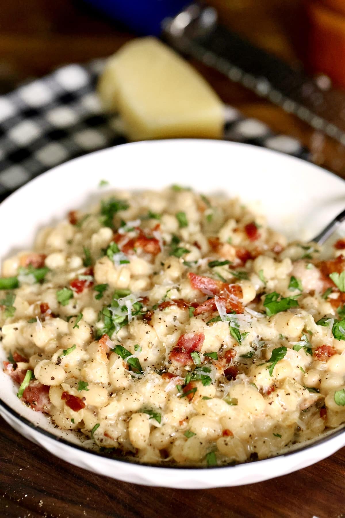 White beans side dish with crispy bacon and parmesan. Bowl of white beans, block of parmesan in background.