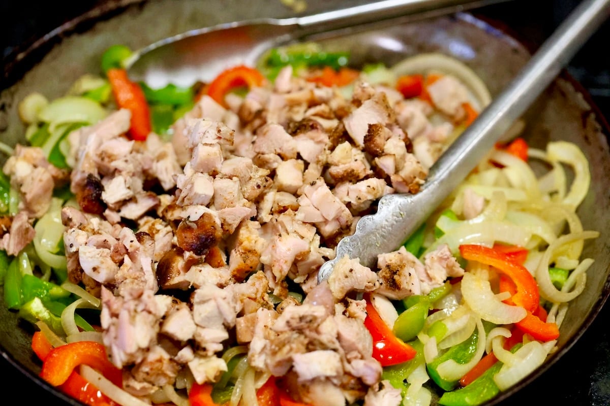 Adding chicken to a skillet with peppers and onions.