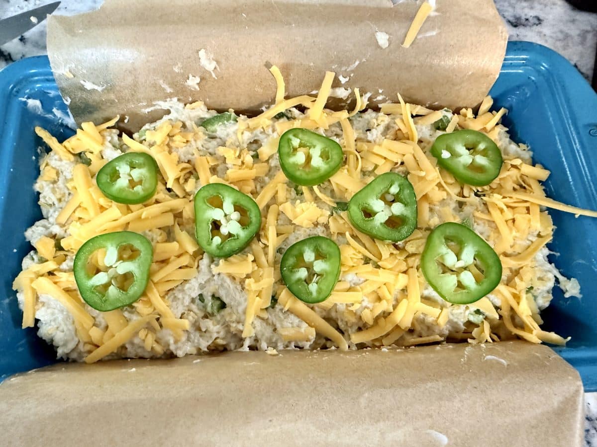 Beer bread in parchment lined bread pan, topped with shredded cheddar and jalapeno rings.