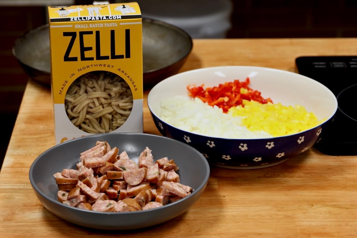 Zelli pasta, diced bell peppers and onions in a bowl, smoked sausage sliced.
